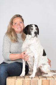 Eine lächelnde Frau sitzt mit einem weißen Hund auf einem Holzpodest.