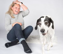 Lachende Frau mit blonden Haaren sitzt neben einem Hund auf einem hellen Hintergrund.